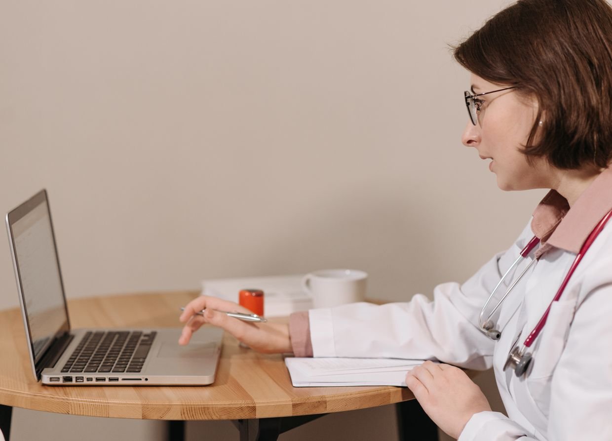 Médica de jaleco trabalhando no notebook em uma mesa de consultório
